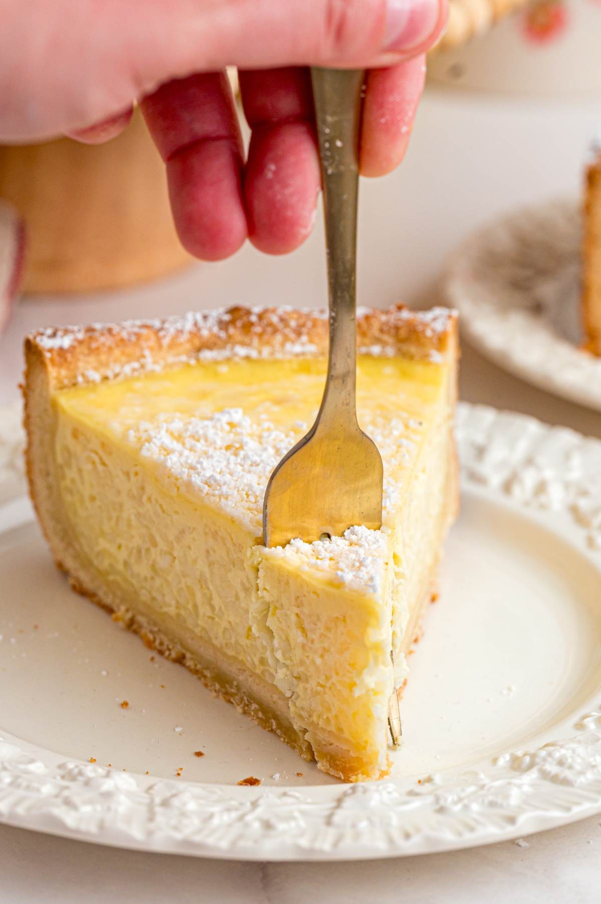 A hand holds a fork, ready to cut into a slice of creamy pie dusted with powdered sugar on a white plate.
