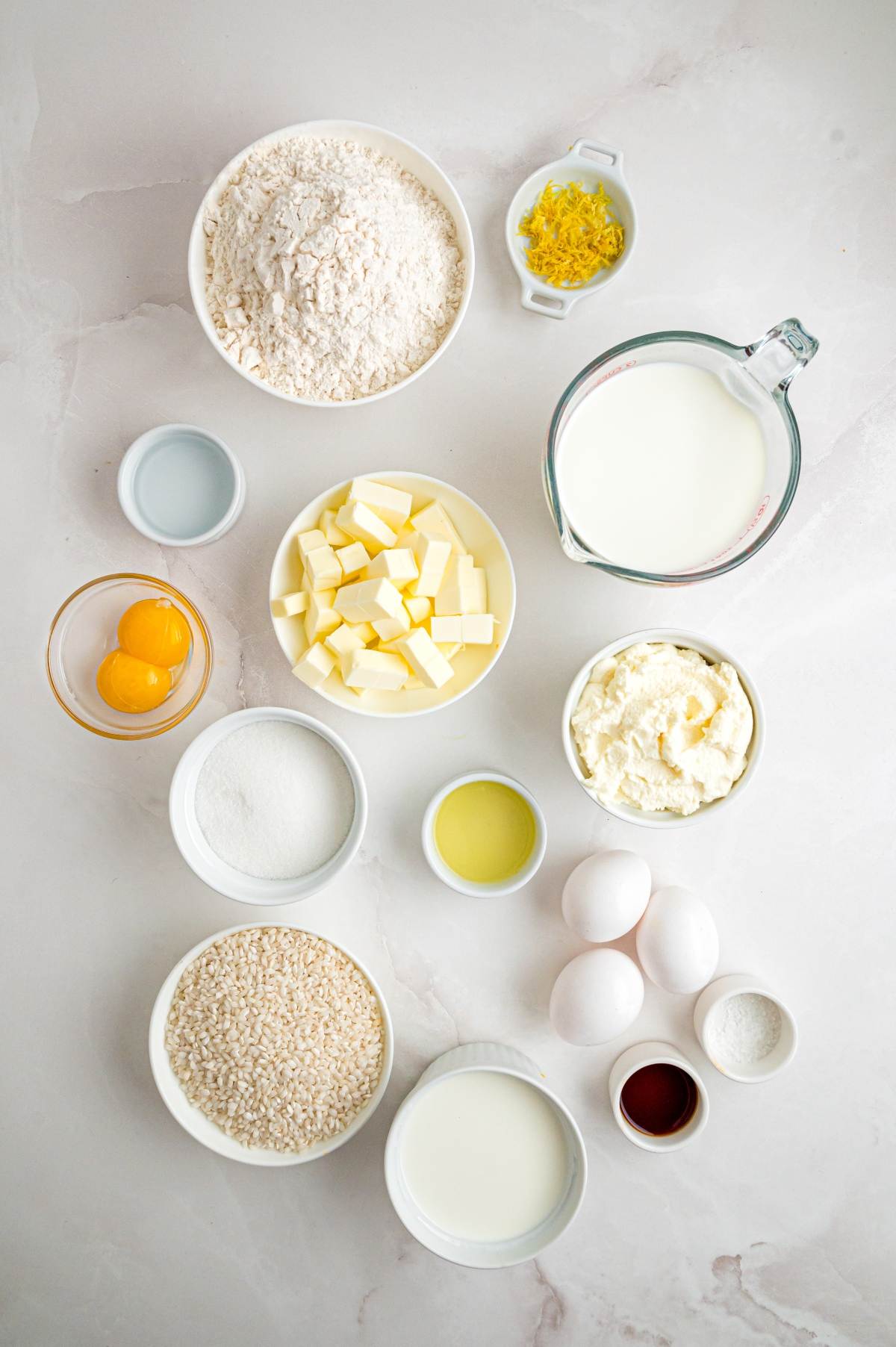 Assorted baking ingredients including flour, butter, sugar, eggs, milk, lemon zest, and vanilla on a white surface.