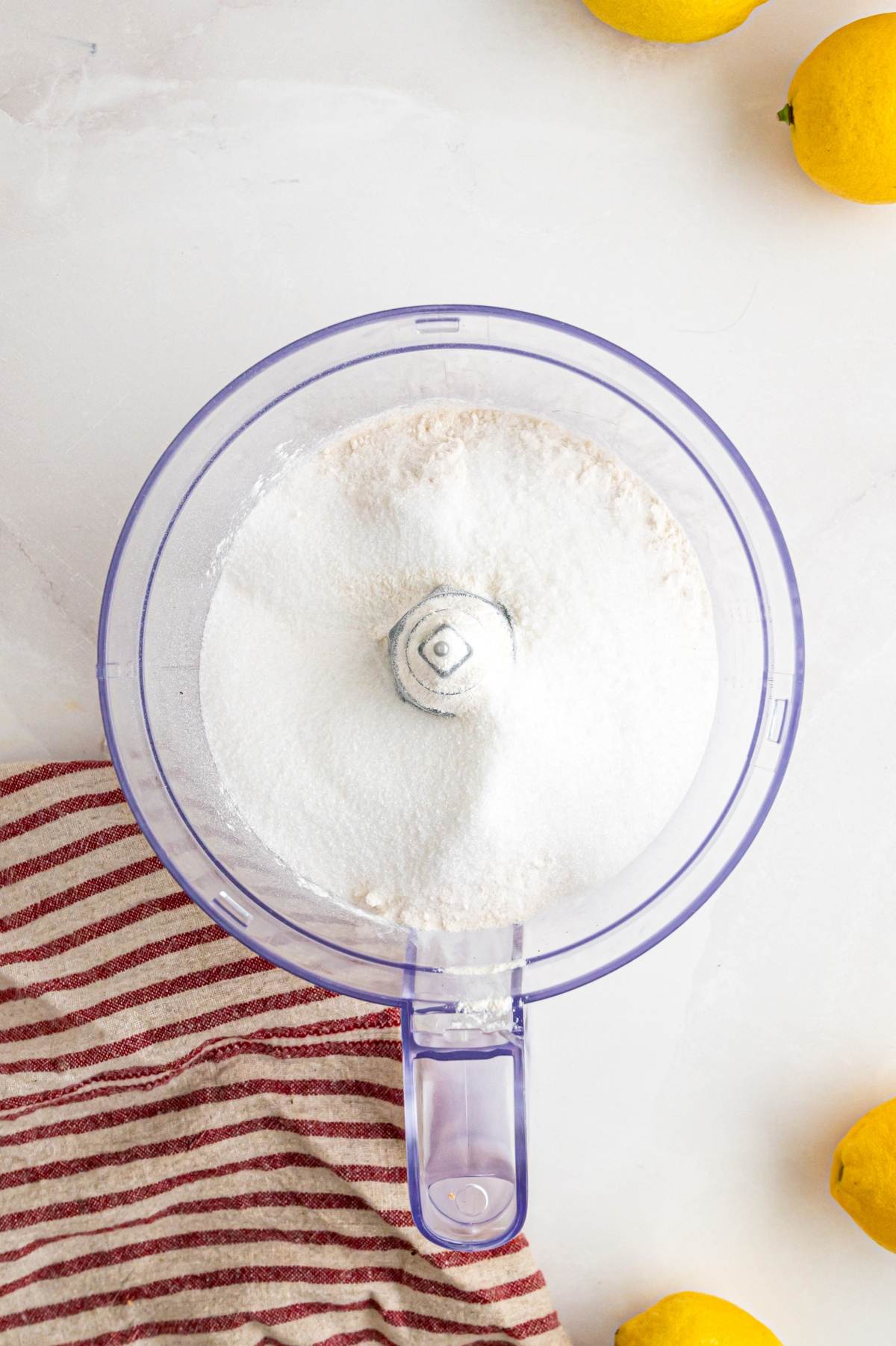 Top view of a food processor with white powder, next to a striped towel and lemons on a light surface.