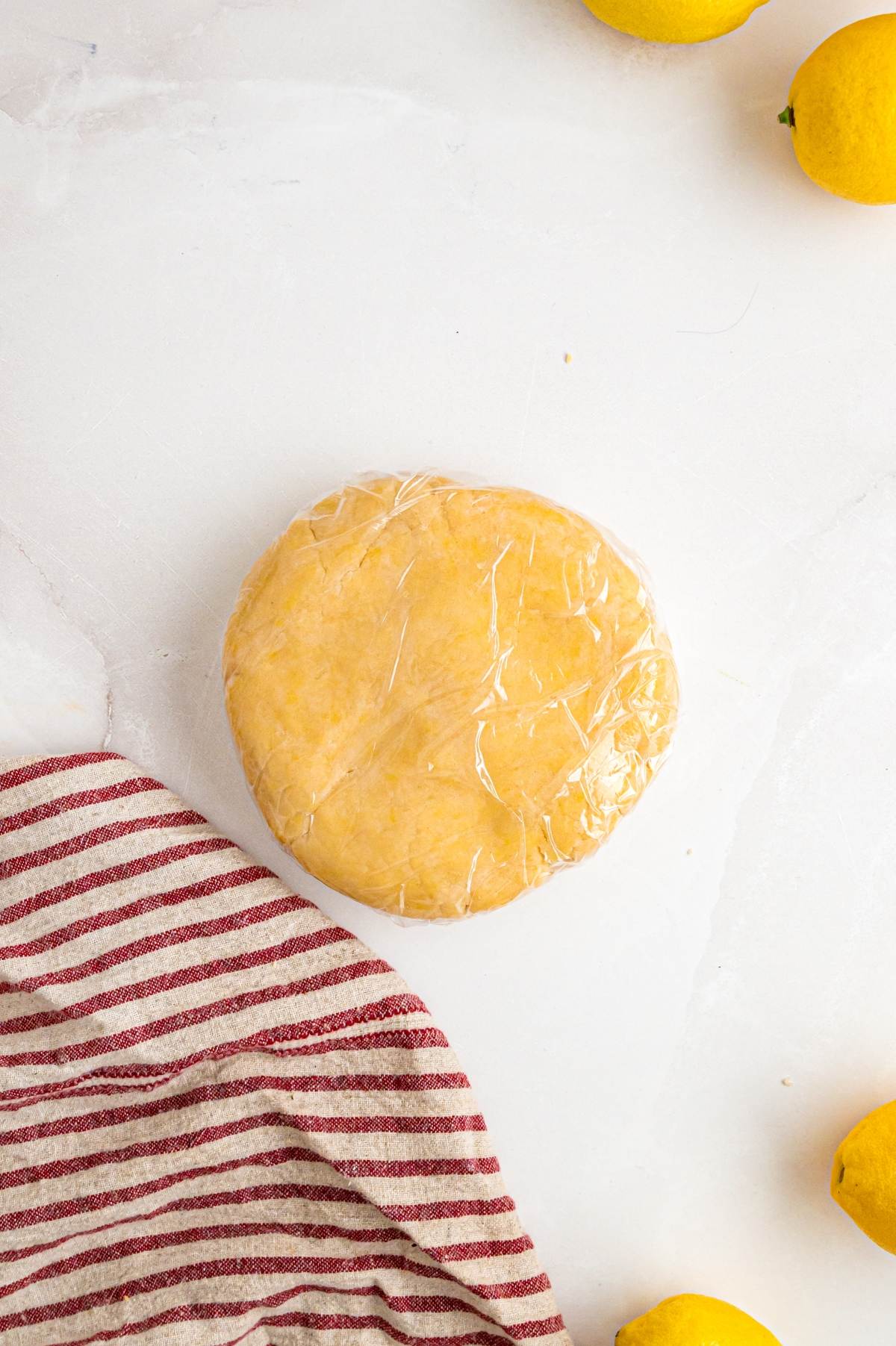 Round disc of dough wrapped in plastic wrap on a white surface with a striped towel and lemons nearby.