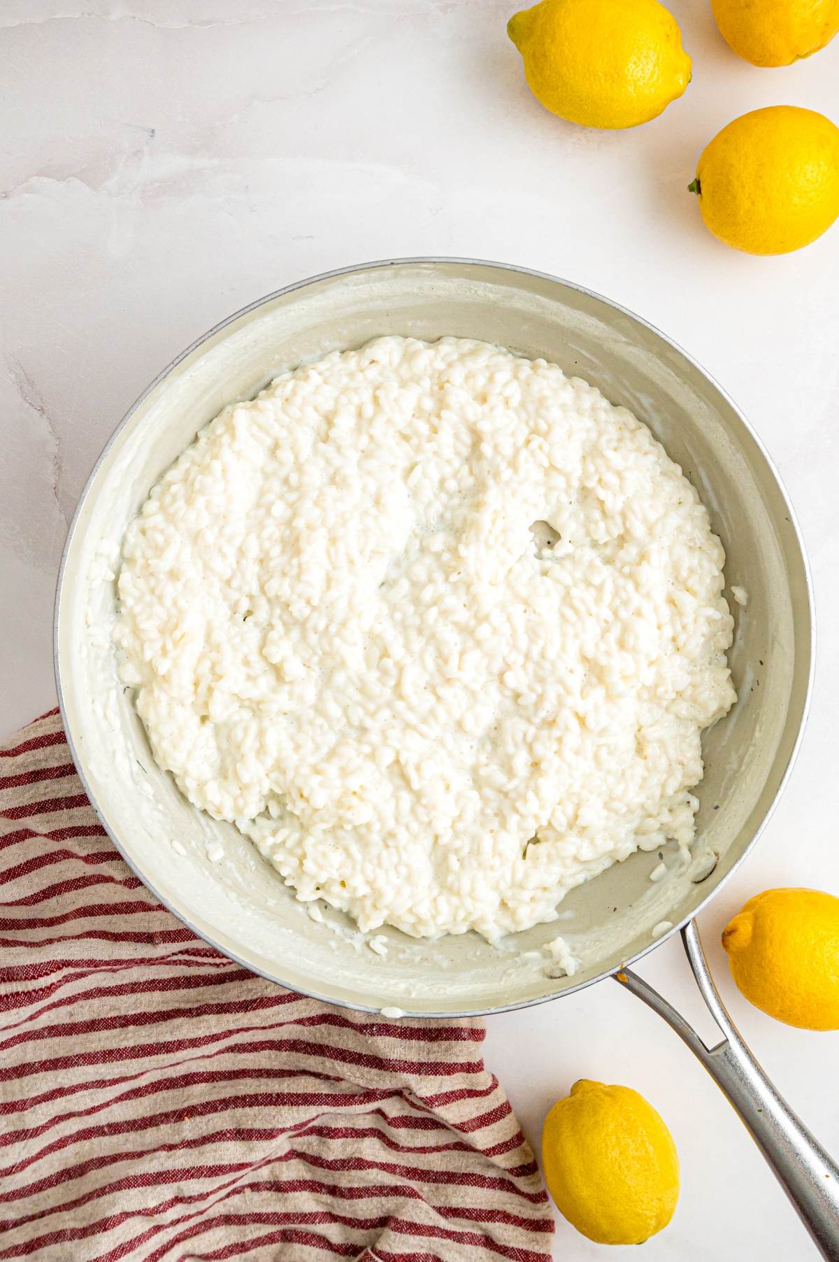 A pan of creamy risotto sits on a striped towel, surrounded by whole lemons on a white surface.