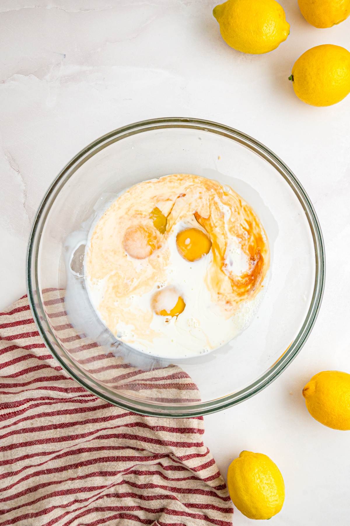 A glass bowl with eggs, milk, and vanilla surrounded by lemons and a red-striped towel on a white surface.