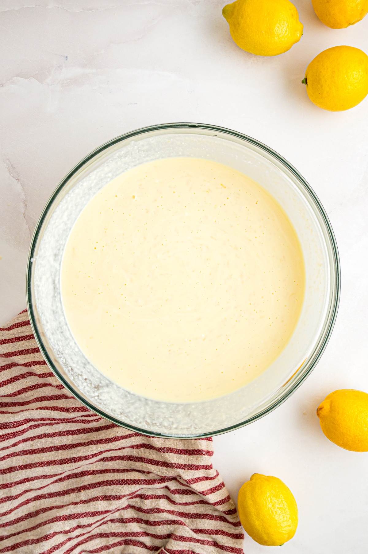 A glass bowl of creamy batter next to whole lemons and a red-striped towel on a white surface.