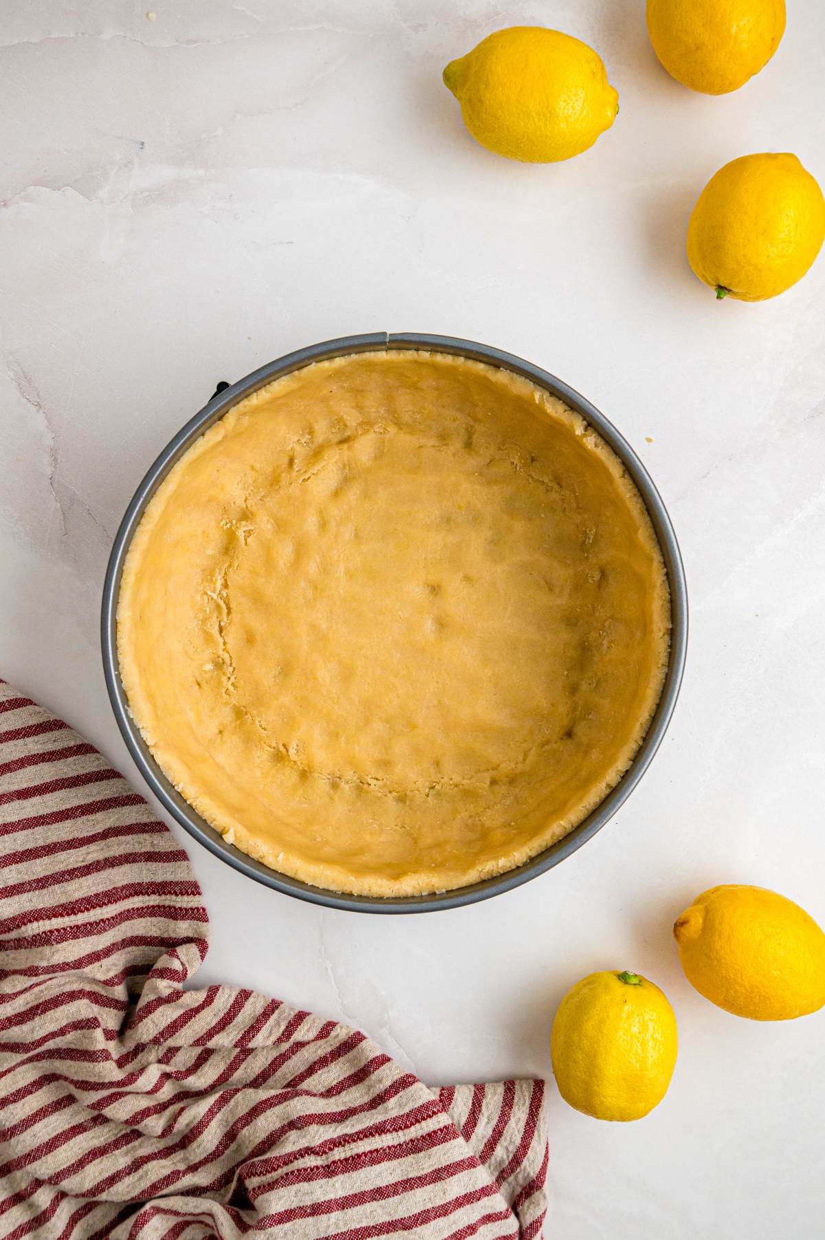 Unbaked pie crust in a round pan, surrounded by lemons and a red-striped kitchen towel on a white surface.