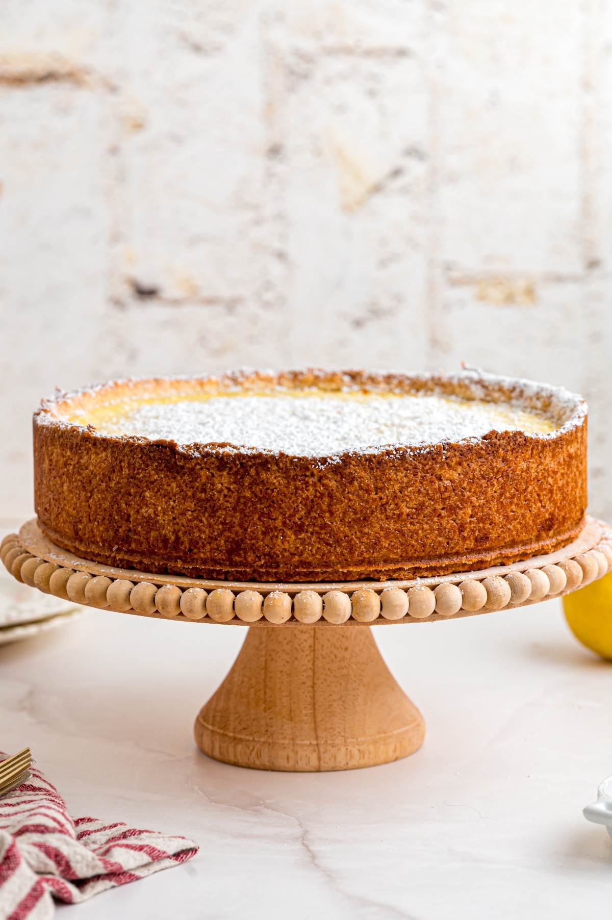 A round cheesecake dusted with powdered sugar sits on a wooden cake stand against a light background.