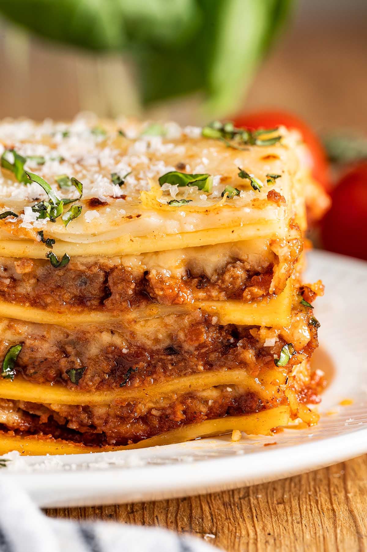 A close-up of a slice of lasagna alla Bolognese with layers of meat, cheese, and pasta, garnished with basil on a white plate.
