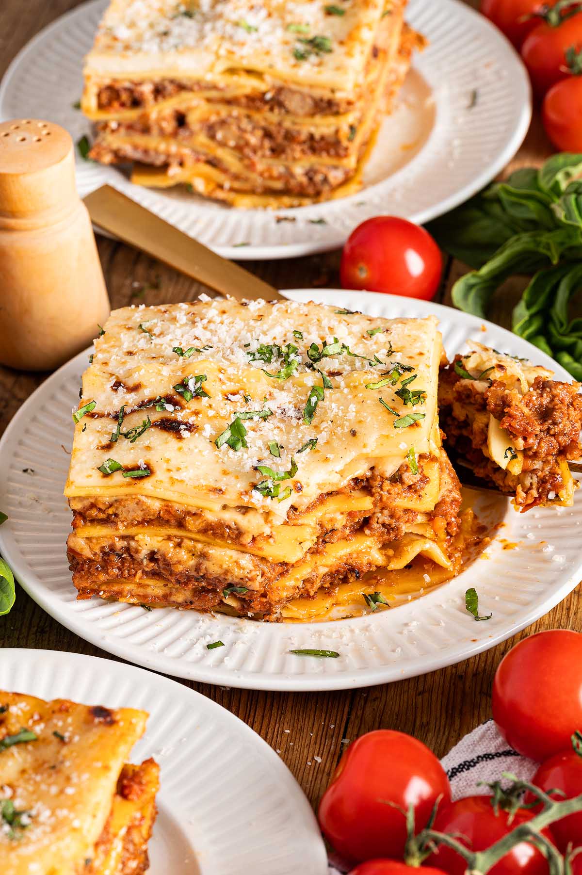 A slice of lasagna alla Bolognese on a plate, topped with grated cheese and herbs, surrounded by fresh tomatoes and basil.