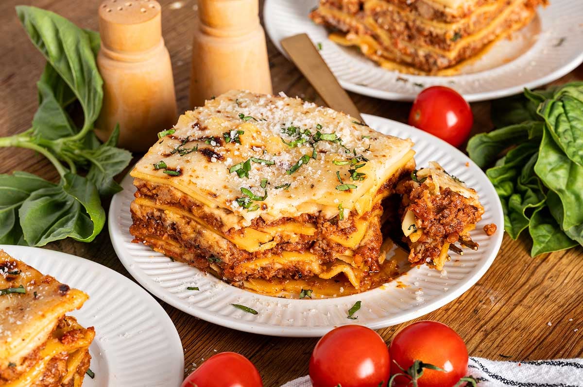 A slice of lasagna alla Bolognese on a plate, garnished with herbs, surrounded by fresh basil and tomatoes.