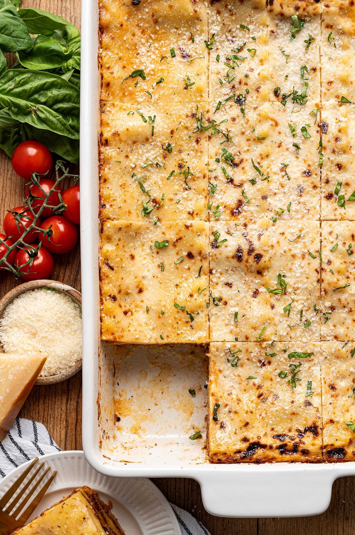 A baked lasagna alla Bolognese in a white dish, sliced into squares with fresh herbs on top and a hearty slice served on a plate nearby.