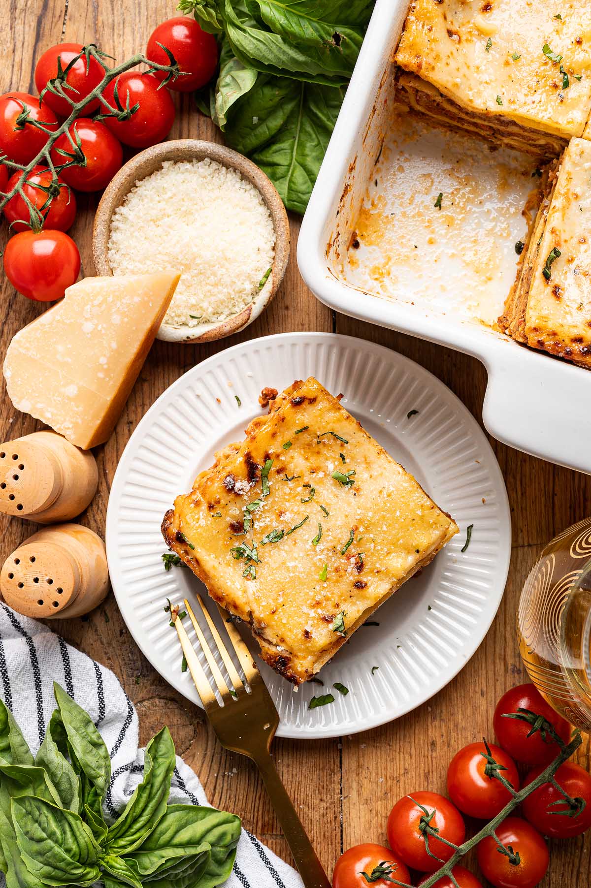 A slice of lasagna alla Bolognese on a plate sits next to parmesan, tomatoes, basil, and a baking dish on a wooden table.