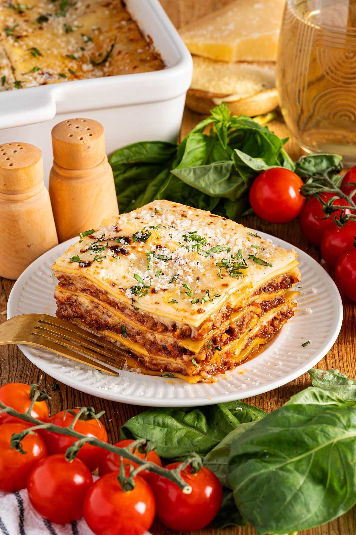 A slice of lasagna alla Bolognese on a plate, surrounded by fresh tomatoes, basil, and a fork.