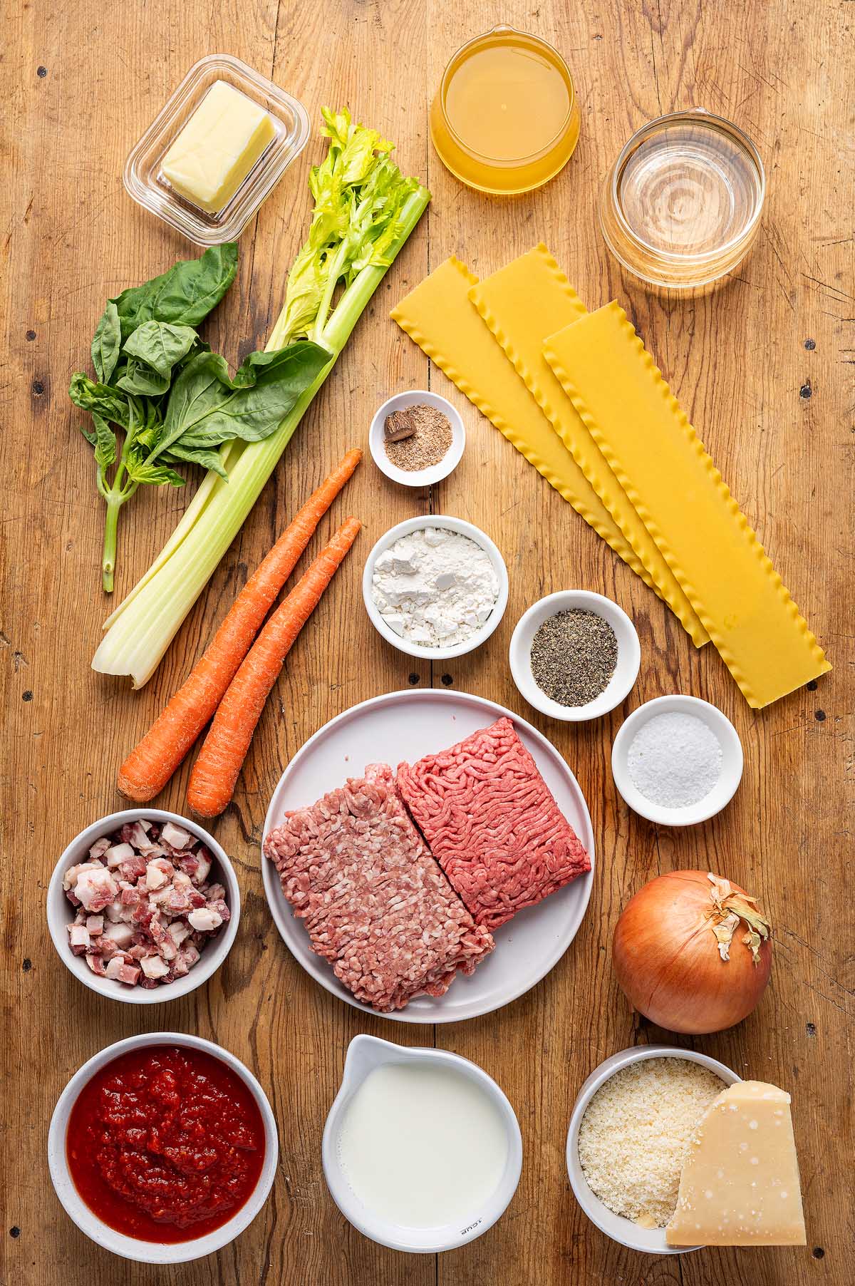 Lasagna alla Bolognese ingredients, such as ground meat, pasta, carrots, celery, onion, cheese, tomato sauce, and seasonings arranged on a table.