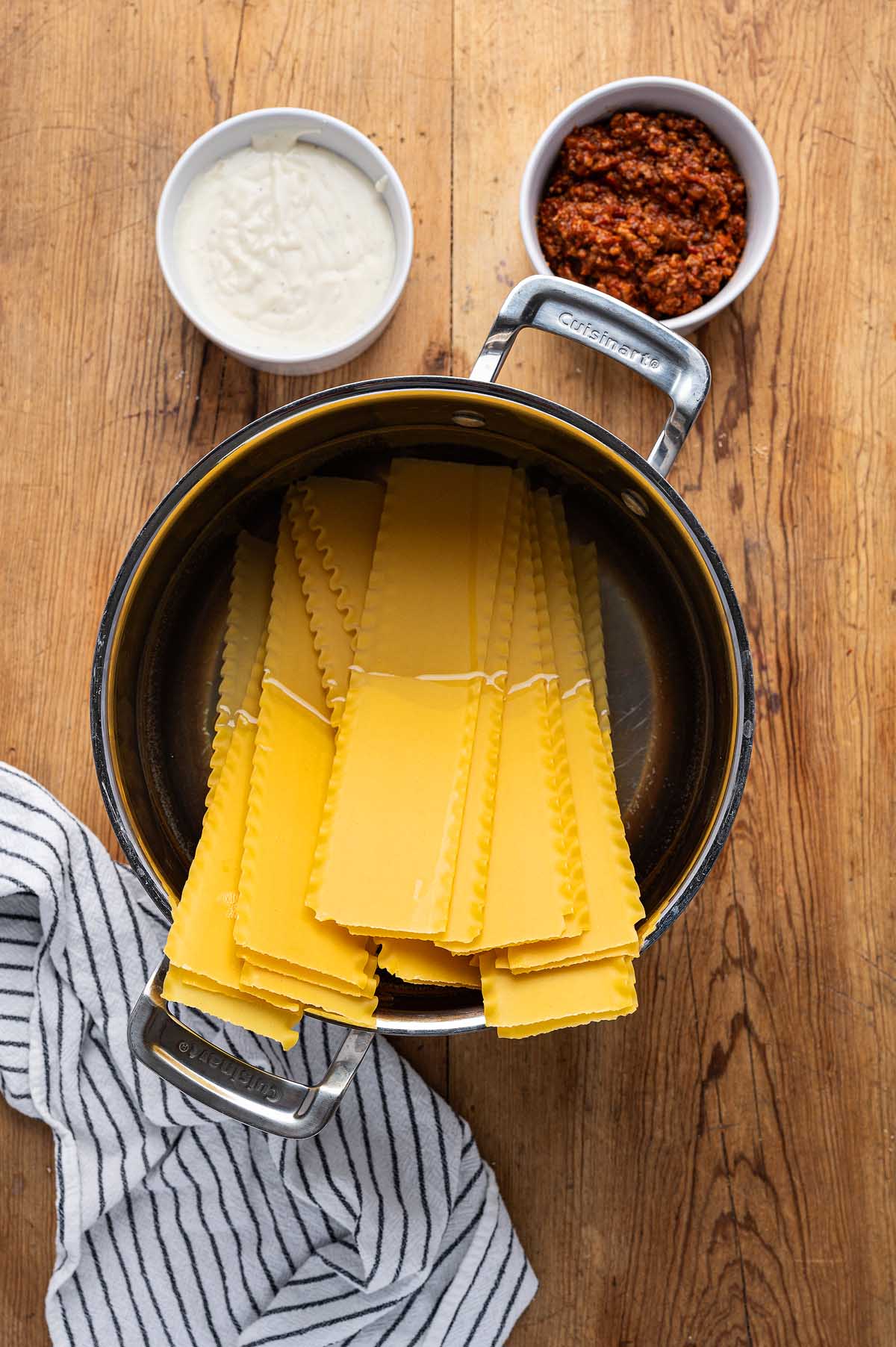 Uncooked lasagna noodles in a pot, with bowls of white sauce and red sauce on a wooden table—everything you need to start your homemade lasagna alla Bolognese.