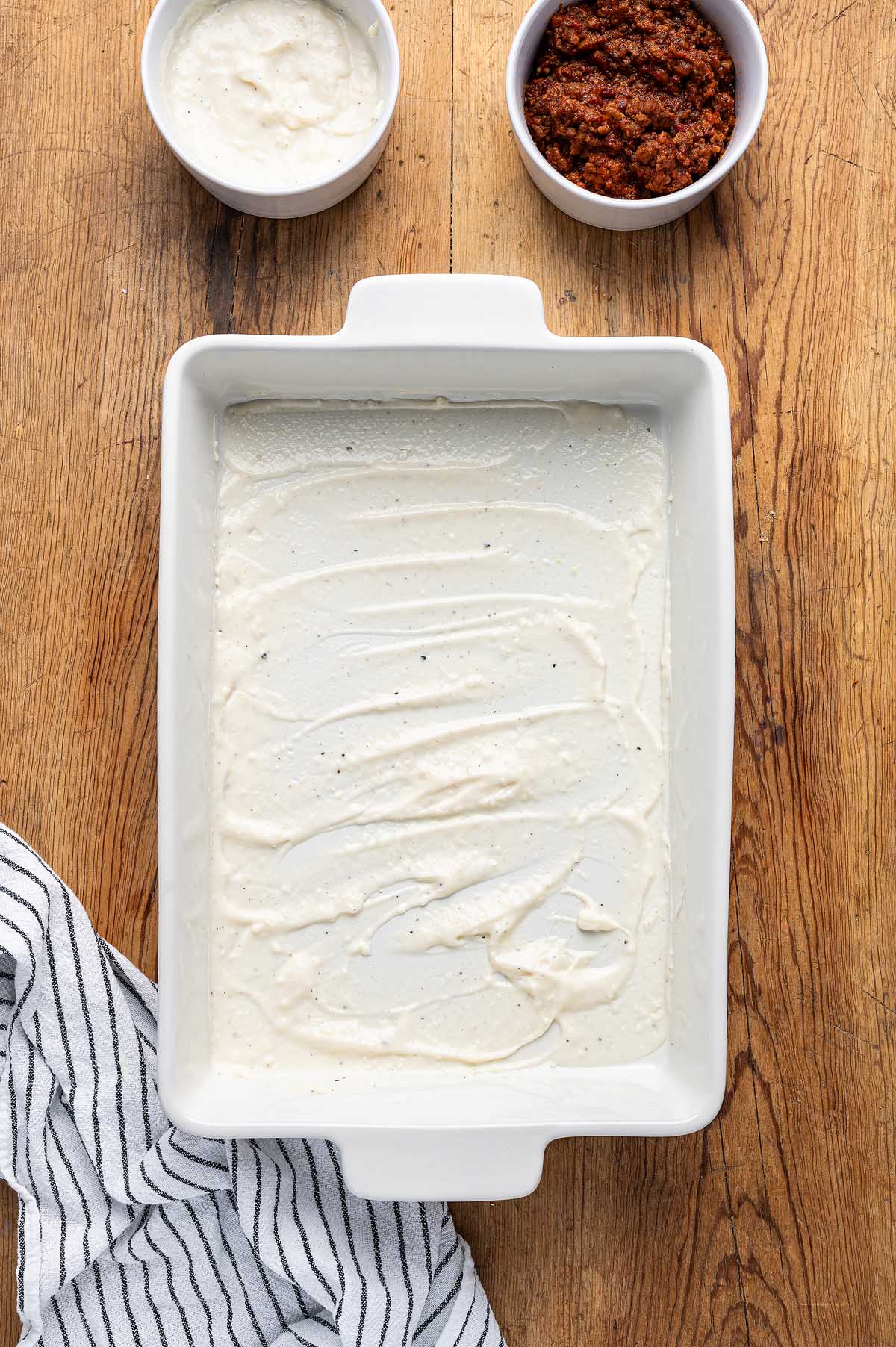 White sauce spread in a rectangular baking dish, ready for layering into classic lasagna alla Bolognese, with bowls of sauce and a striped towel beside it on a wooden table.