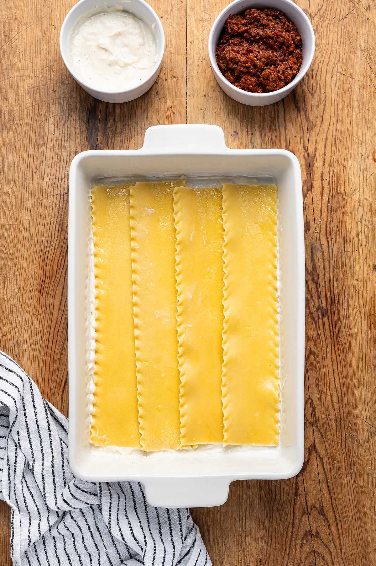 Four lasagna noodles in a white baking dish, with bowls of white sauce and red sauce on a wooden surface—perfect for making classic lasagna alla Bolognese.