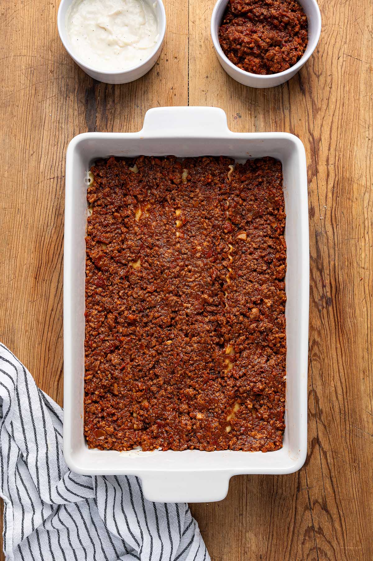 A rectangular dish of lasagna alla Bolognese with meat sauce sits next to bowls of sauce and ricotta on a wooden table.