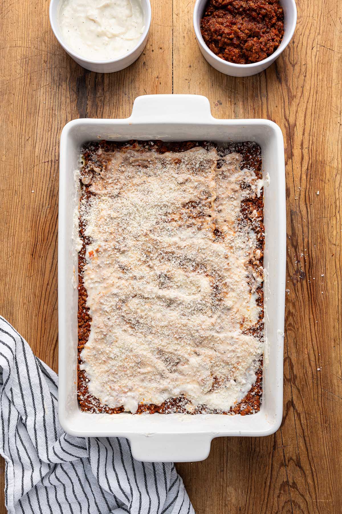 Rectangular baking dish with lasagna alla Bolognese being assembled, next to bowls of sauce and a striped kitchen towel.