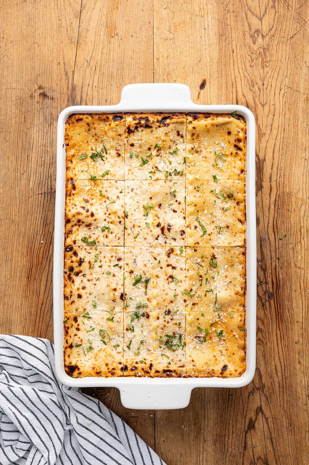 Baked lasagna alla Bolognese in a white dish, cut into squares, topped with fresh herbs and melted cheese on a wooden table.
