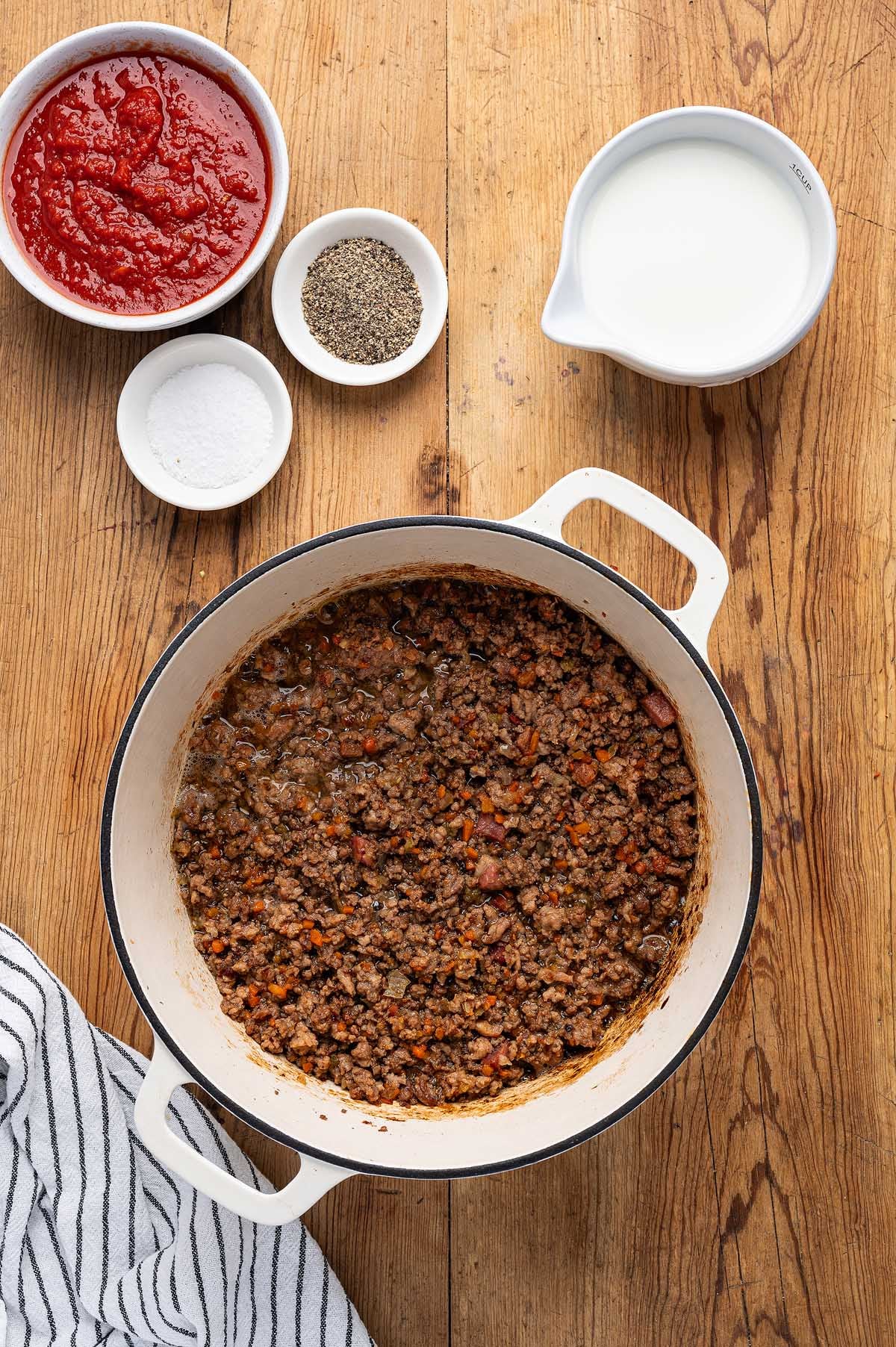 A pot of cooked ground meat sits beside bowls of tomato sauce, spices, and milk on a wooden surface—classic ingredients for lasagna alla Bolognese.