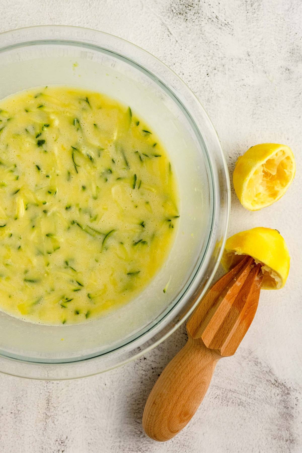 A glass bowl with grated zucchini mixture sits beside squeezed lemon halves and a wooden citrus reamer—perfect for preparing homemade lemon zucchini bread.