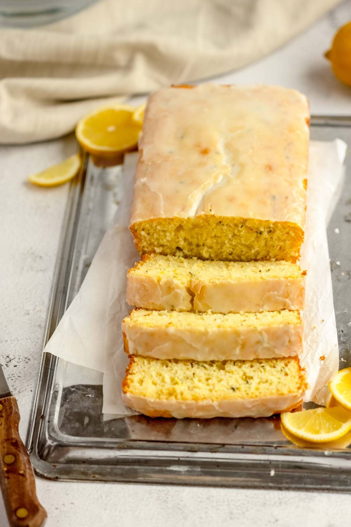 A glazed lemon zucchini bread loaf cake with three slices cut, on parchment paper, surrounded by lemon wedges.