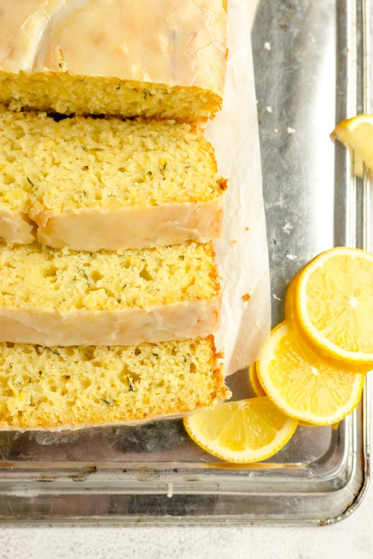 Sliced lemon zucchini bread loaf with glaze on parchment paper, garnished with fresh lemon slices.