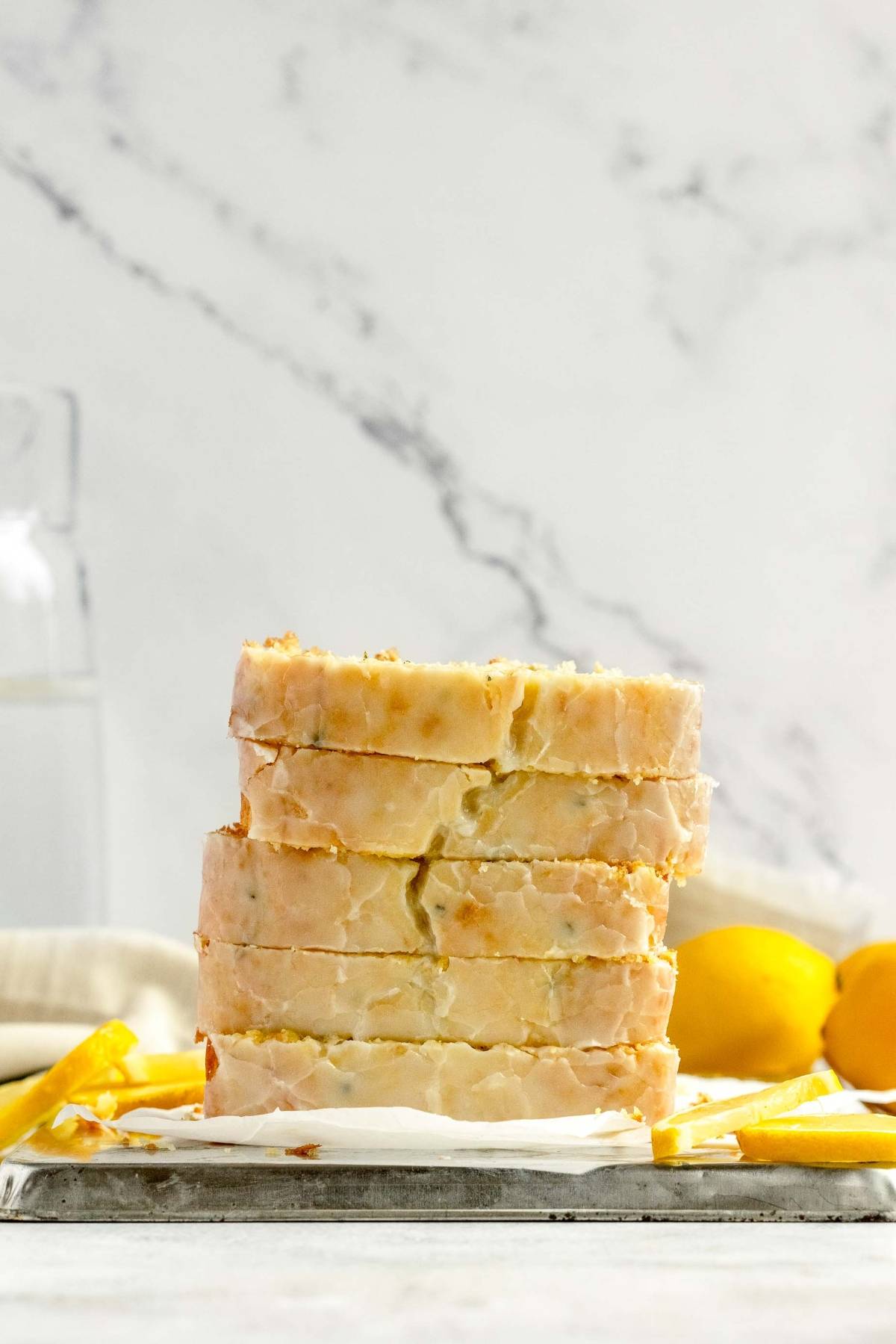 Five slices of glazed lemon zucchini bread stacked on a tray, with lemons and a glass of water in the background.