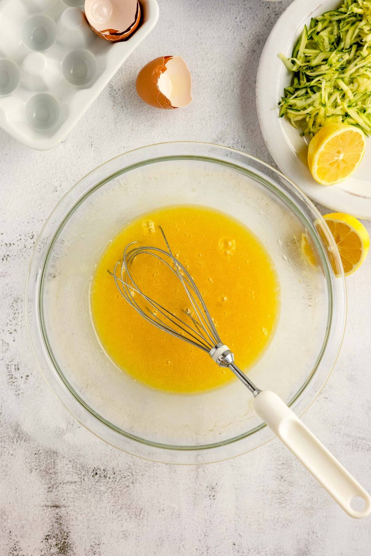 A whisk in a bowl of beaten eggs sits beside eggshells, grated zucchini, and a lemon on a white surface—perfect ingredients for making fresh lemon zucchini bread.