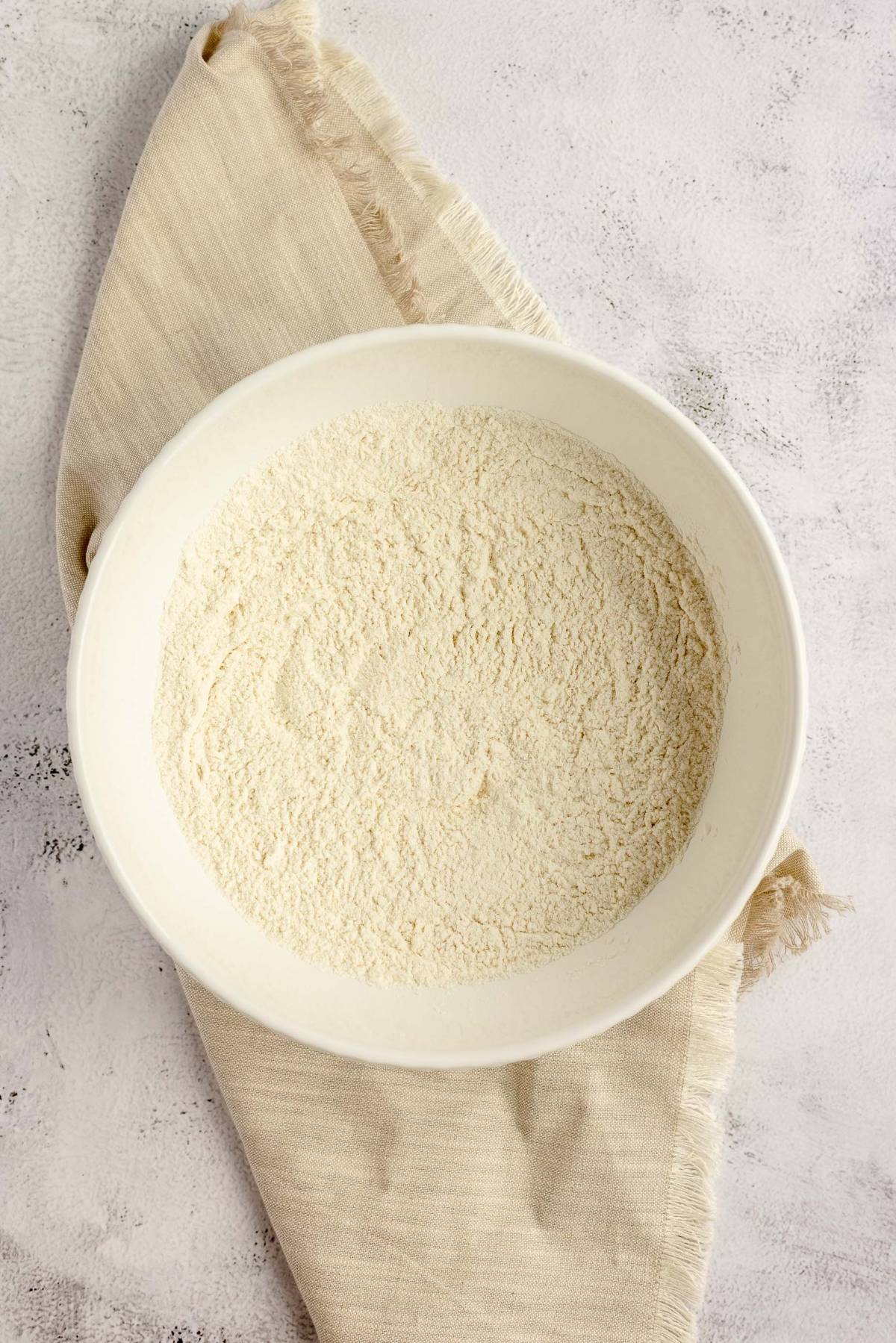 A white bowl filled with flour sits on a beige cloth against a light textured background, ready to be used for baking lemon zucchini bread.