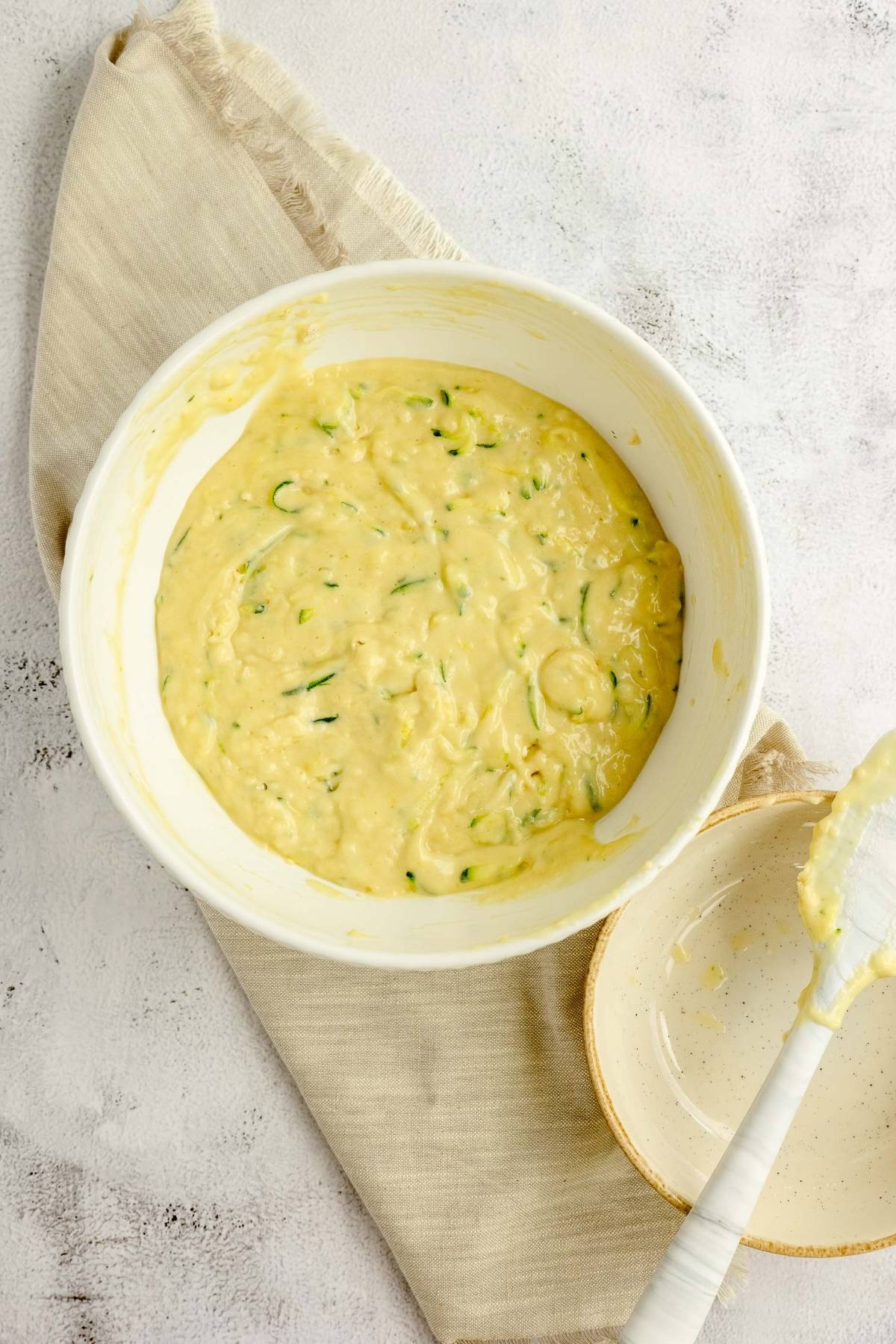 Bowl of lemon zucchini bread batter with a spatula on a beige cloth and a small empty bowl nearby.
