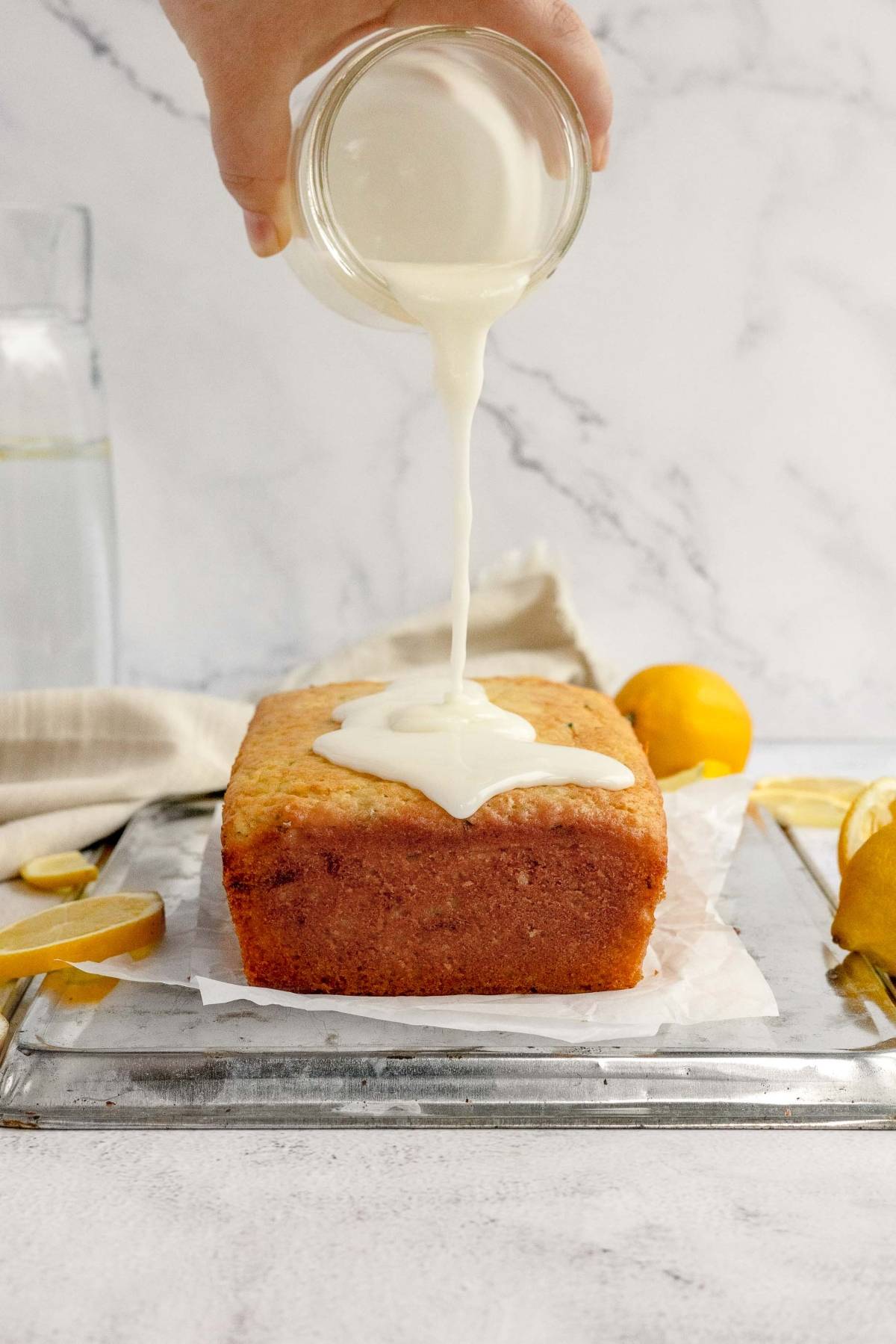 A hand pours white icing over a loaf of lemon zucchini bread on a wire rack, with fresh lemons nearby.