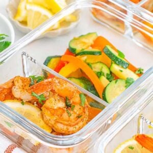 Glass containers with Meal Prep Cajun Shrimp and Veggies, accompanied by lemon slices and mixed sautéed vegetables, arranged on a white surface.