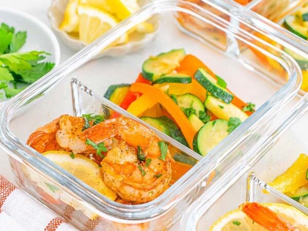 Glass containers with Meal Prep Cajun Shrimp and Veggies, accompanied by lemon slices and mixed sautéed vegetables, arranged on a white surface.