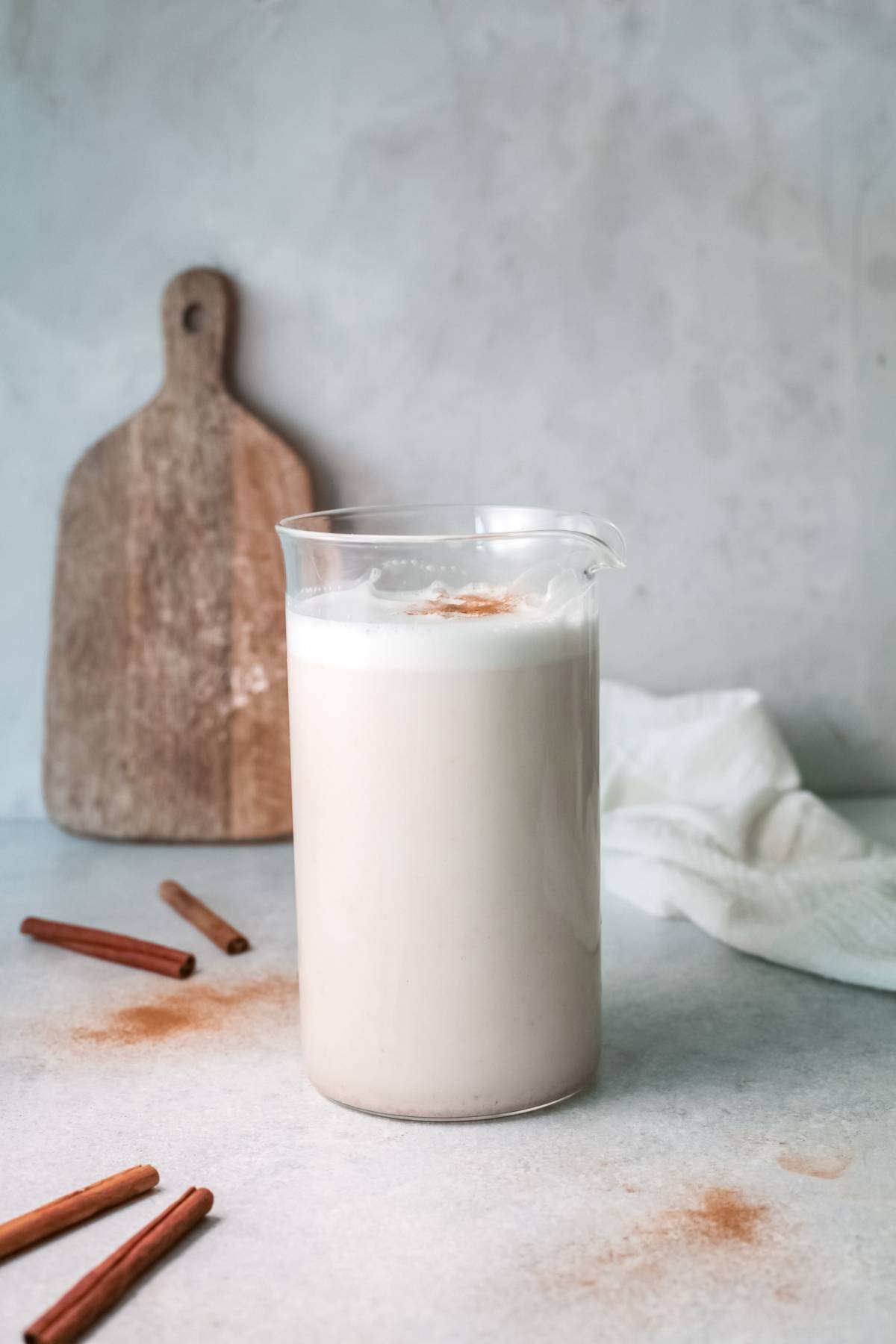 A glass pitcher of homemade horchata topped with cinnamon, with cinnamon sticks and a wooden board in the background—perfect for learning how to make this refreshing iced rice milk beverage.