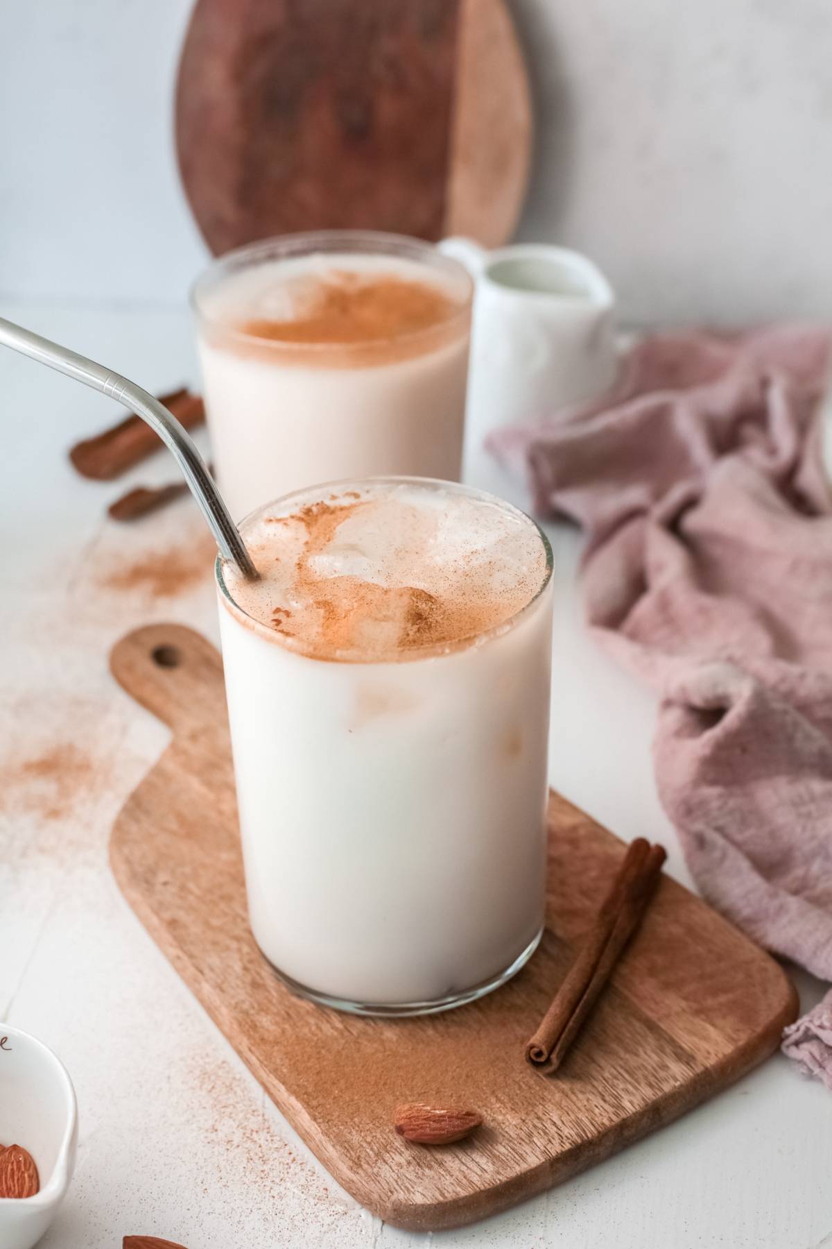 Two glasses of Mexican Horchata, an iced rice milk beverage topped with cinnamon and served with metal straws, sit on a wooden board next to a cinnamon stick.