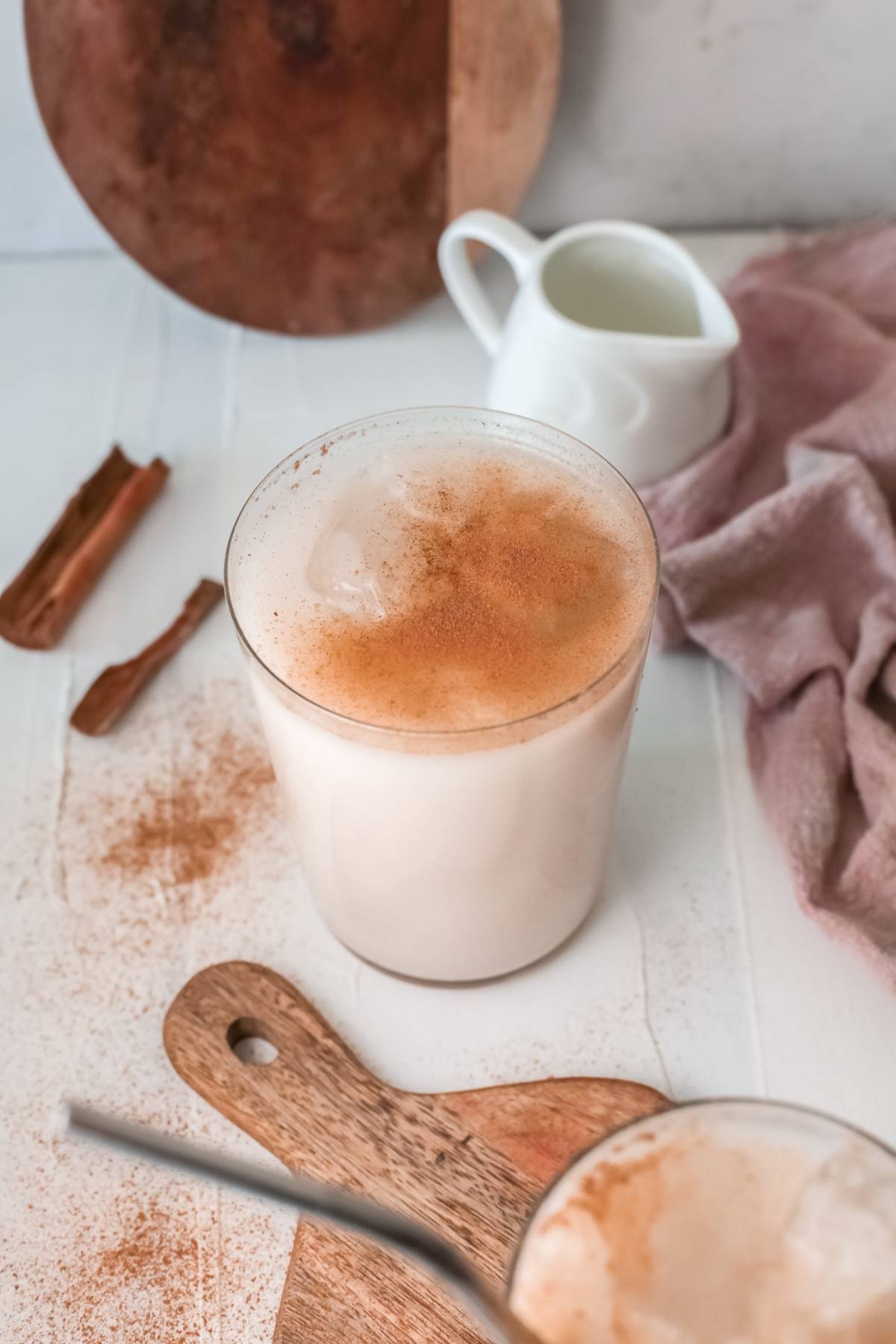 A glass of iced rice milk beverage horchata with cinnamon on top sits next to a cinnamon stick and a white creamer, showcasing this traditional Mexican drink recipe on a table.