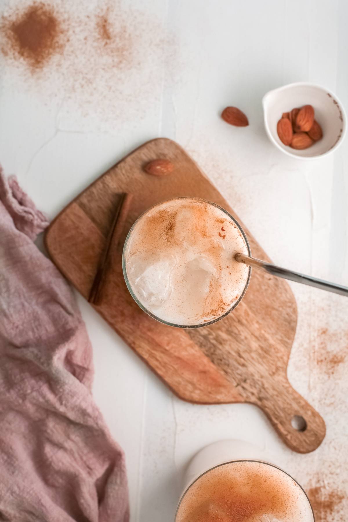Iced latte with cinnamon on a wooden board, beside almonds and a pink cloth on a white surface, inspired by the classic homemade horchata iced rice milk beverage.