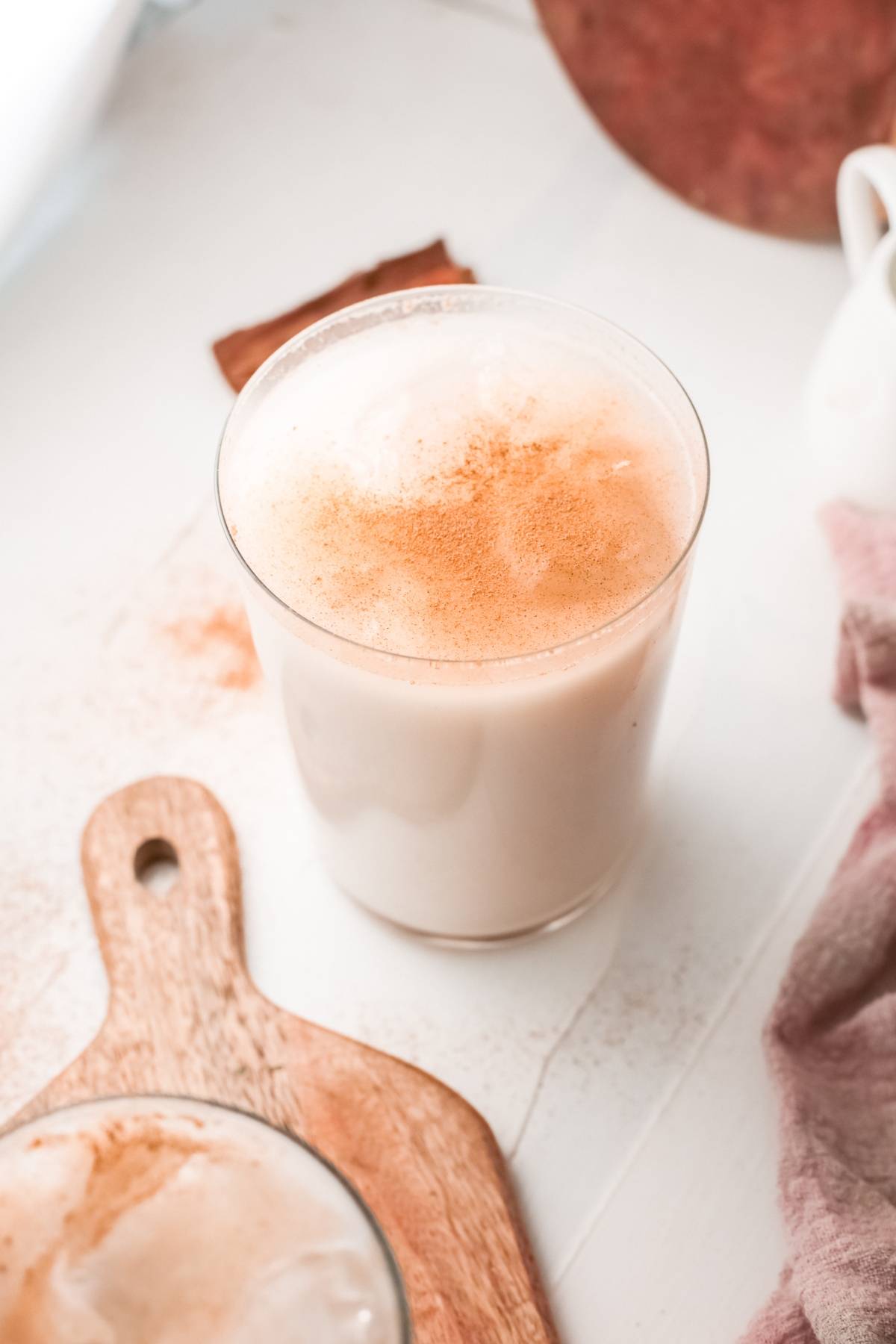 A glass of frothy, creamy Mexican Horchata sits on a white surface next to a wooden board, topped with cinnamon for the perfect iced rice milk beverage.