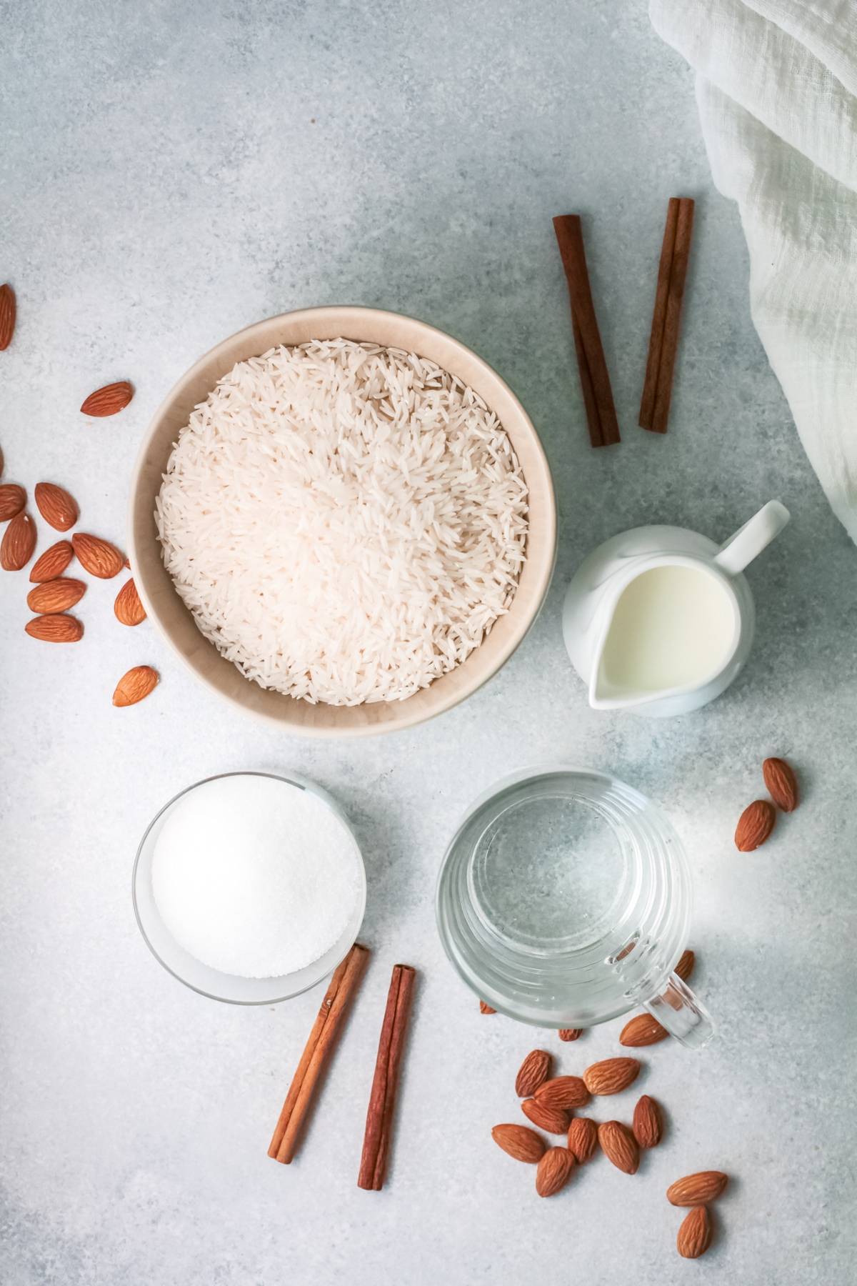 Bowl of rice, milk, sugar, water, almonds, and cinnamon sticks on a light surface—everything you need to learn how to make horchata or enjoy a refreshing iced rice milk beverage at home.