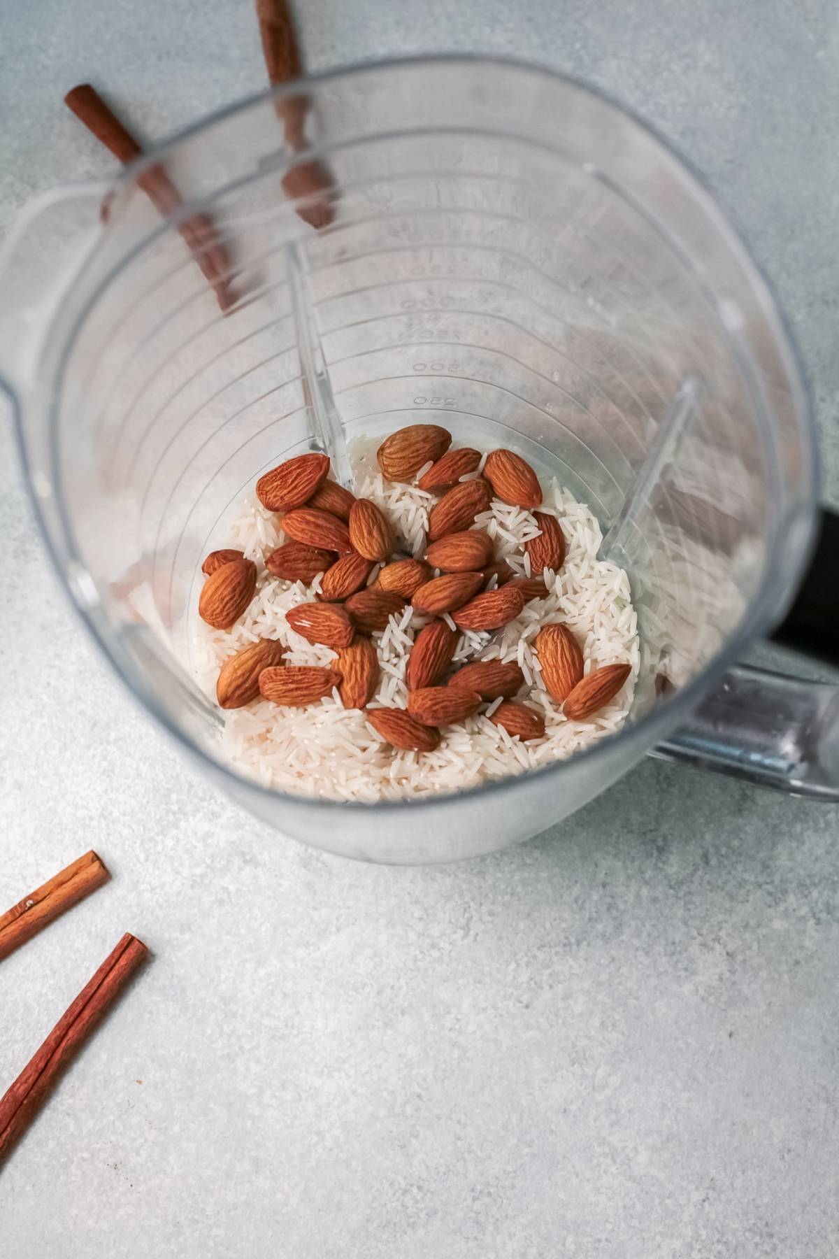 Overhead view of a blender with raw rice and whole almonds inside, next to cinnamon sticks on a light surface—perfect for preparing homemade horchata, the traditional Mexican drink recipe for iced rice milk beverage.