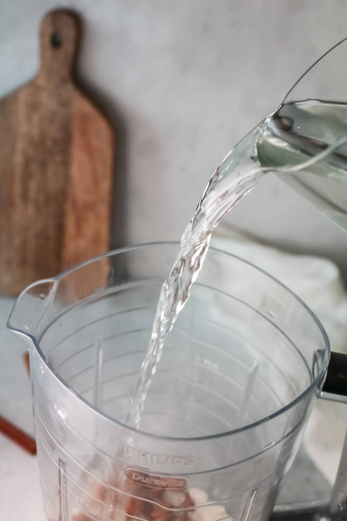 Water being poured from a glass pitcher into a clear blender jar, ready to show how to make horchata—a traditional Mexican drink recipe—set against a wooden cutting board in the background.