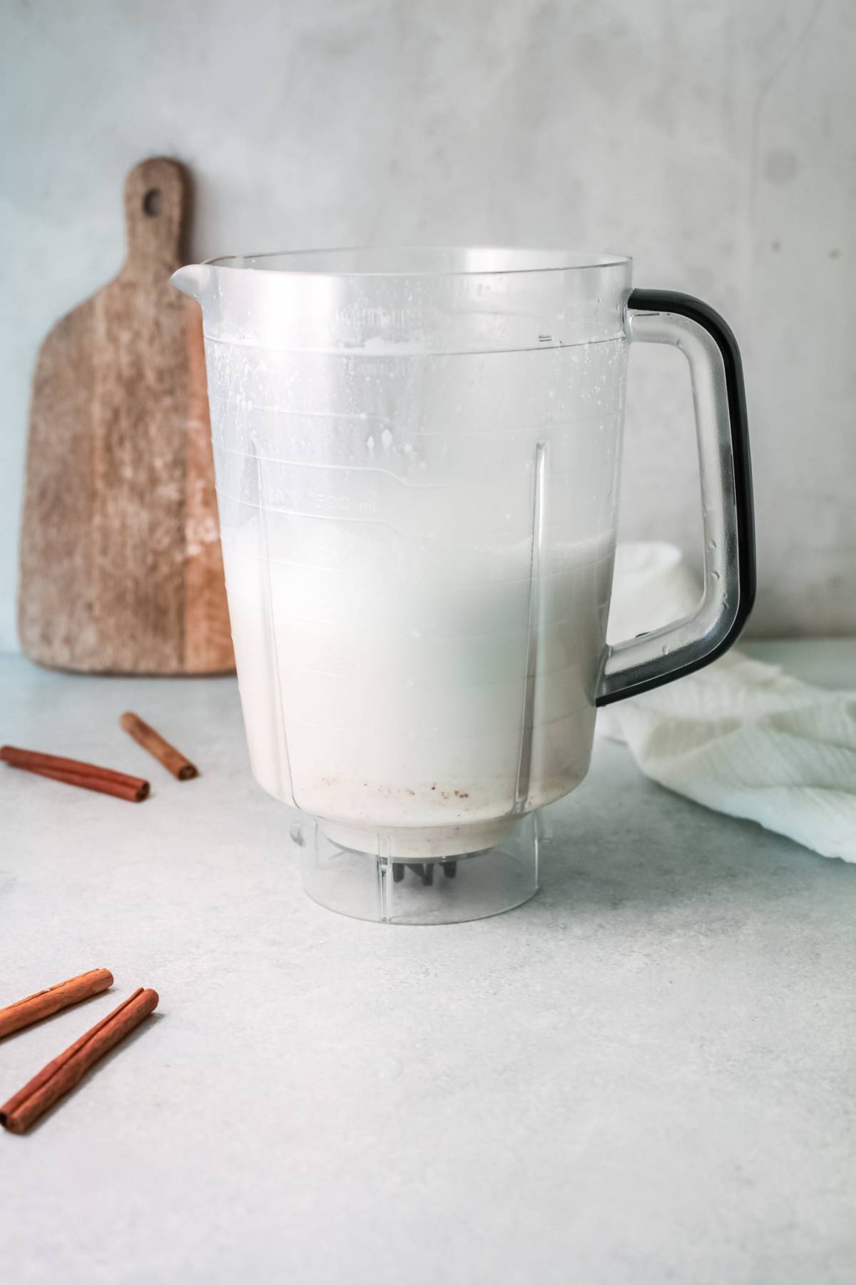 A blender pitcher filled with milk sits beside cinnamon sticks and a wooden board on a countertop, perfect for preparing a traditional Mexican drink recipe like homemade Mexican Horchata.
