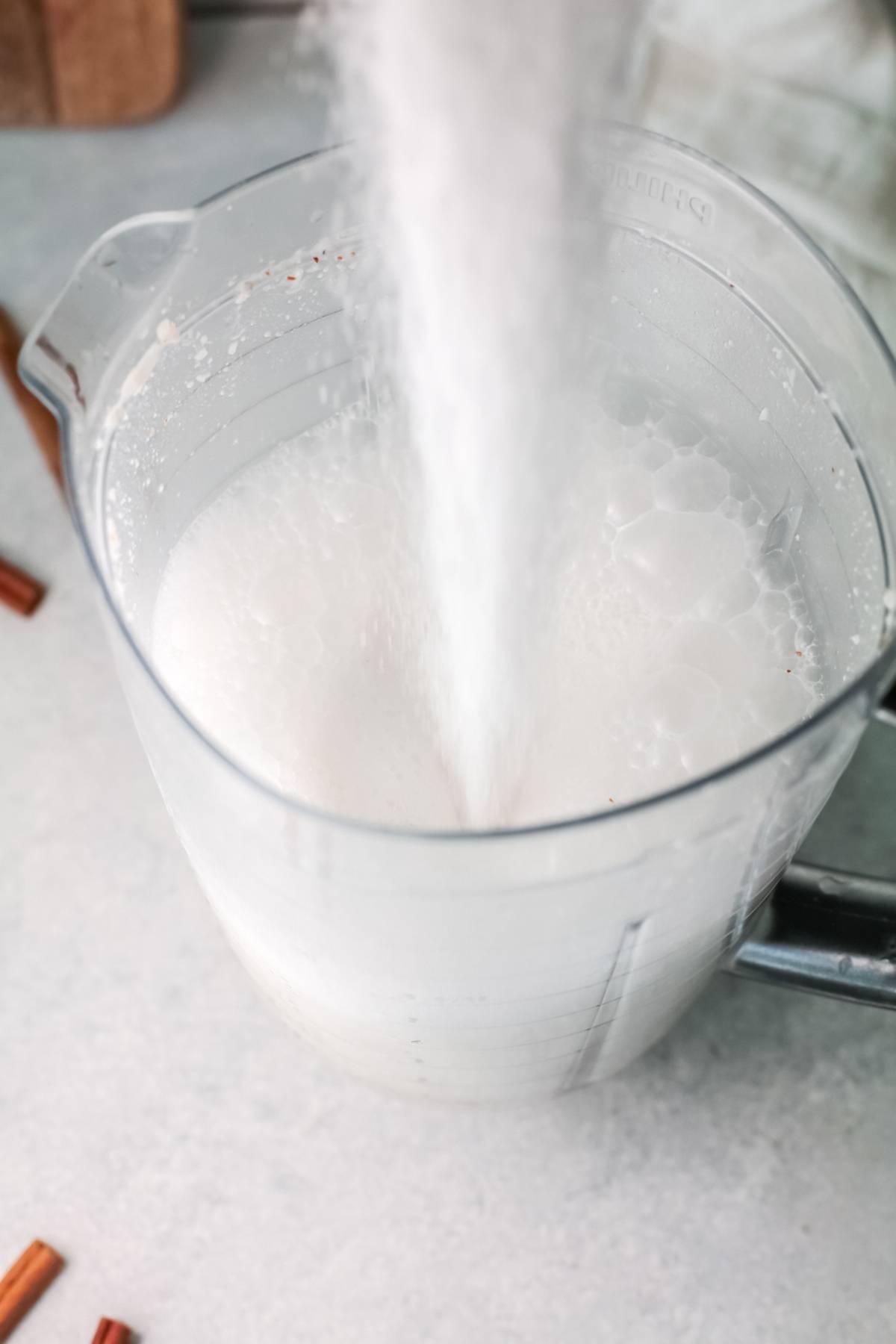 Sugar being poured into a large clear measuring jug filled with a milky liquid on a light surface, perfectly capturing how to make horchata—an authentic iced rice milk beverage enjoyed in Mexican Horchata recipes.