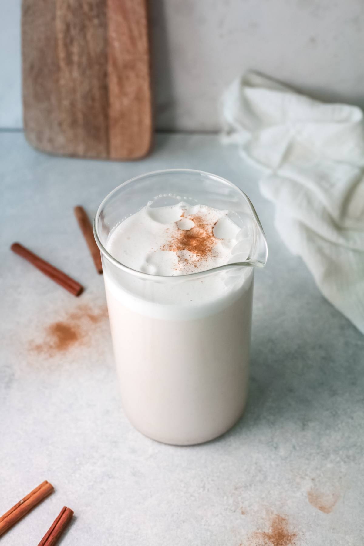 A glass pitcher of frothy homemade horchata topped with cinnamon, surrounded by cinnamon sticks on a light surface—a classic traditional Mexican drink recipe.