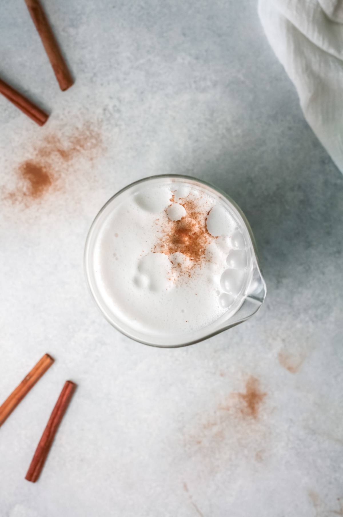 A glass of frothy iced rice milk beverage topped with cinnamon, surrounded by cinnamon sticks on a light surface—a refreshing take on the traditional Mexican drink recipe.