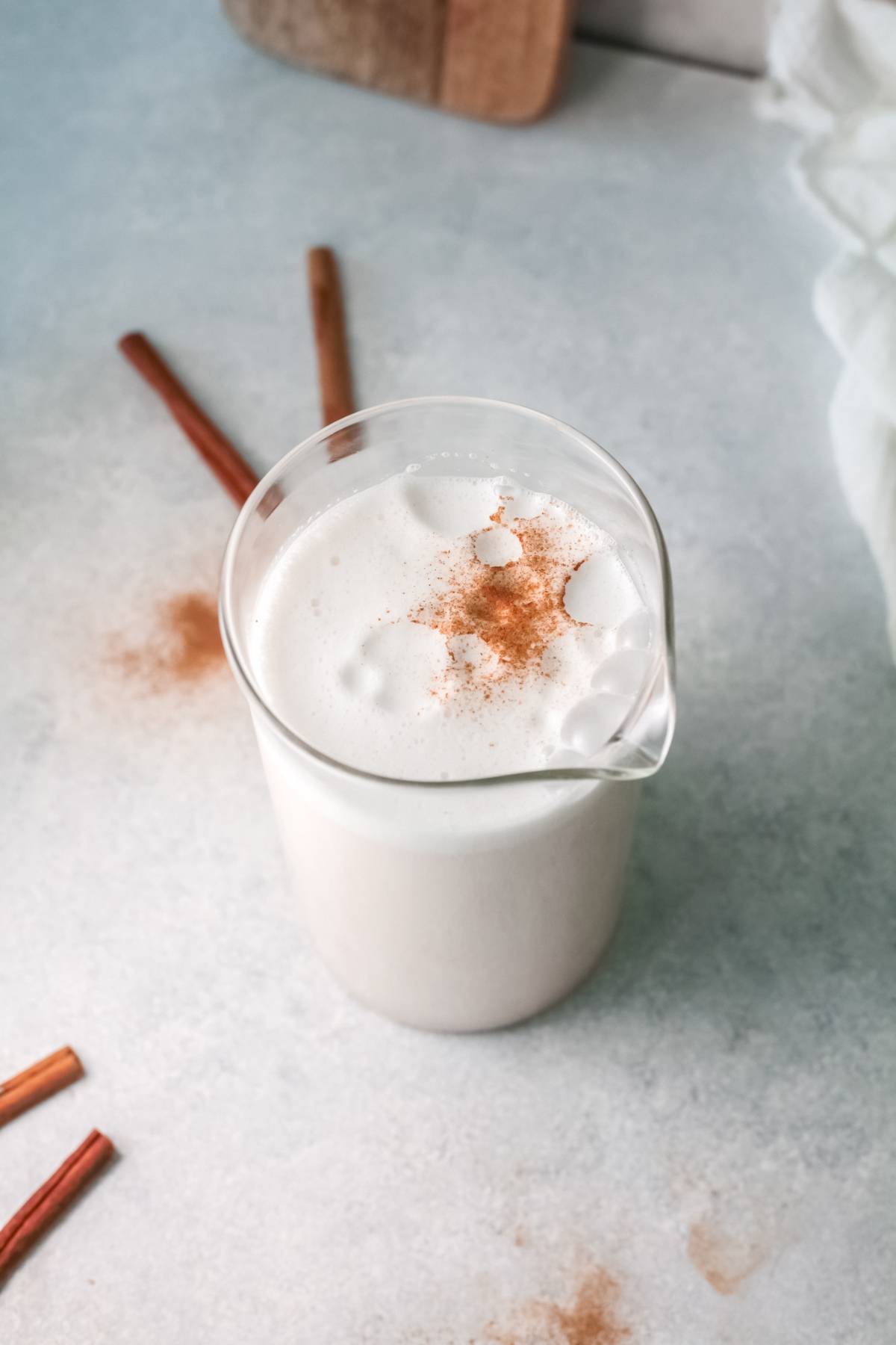 A glass pitcher of frothy Mexican Horchata, an iced rice milk beverage, topped with cinnamon and surrounded by cinnamon sticks on a light surface.