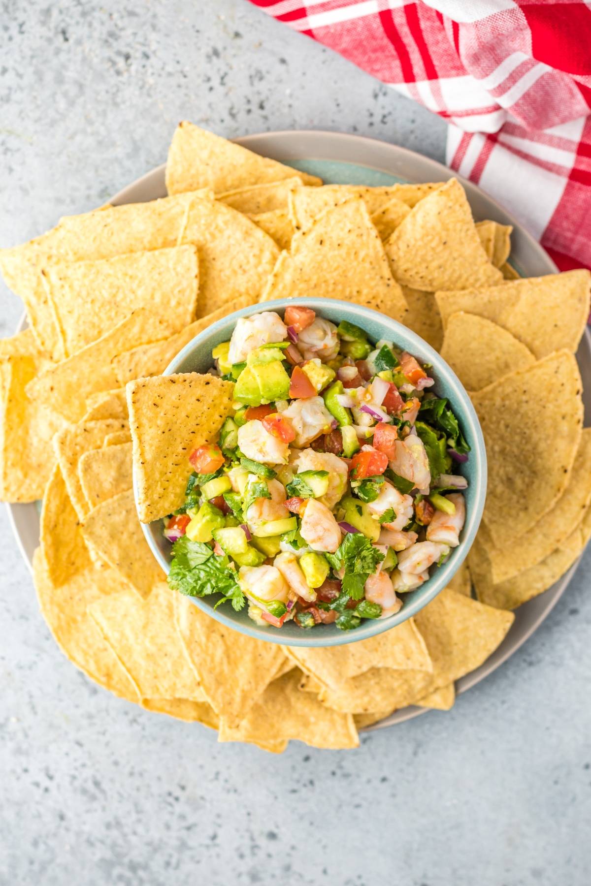 A bowl of avocado shrimp salsa inspired by Mexican ceviche sits surrounded by tortilla chips on a plate, with a red and white napkin nearby.