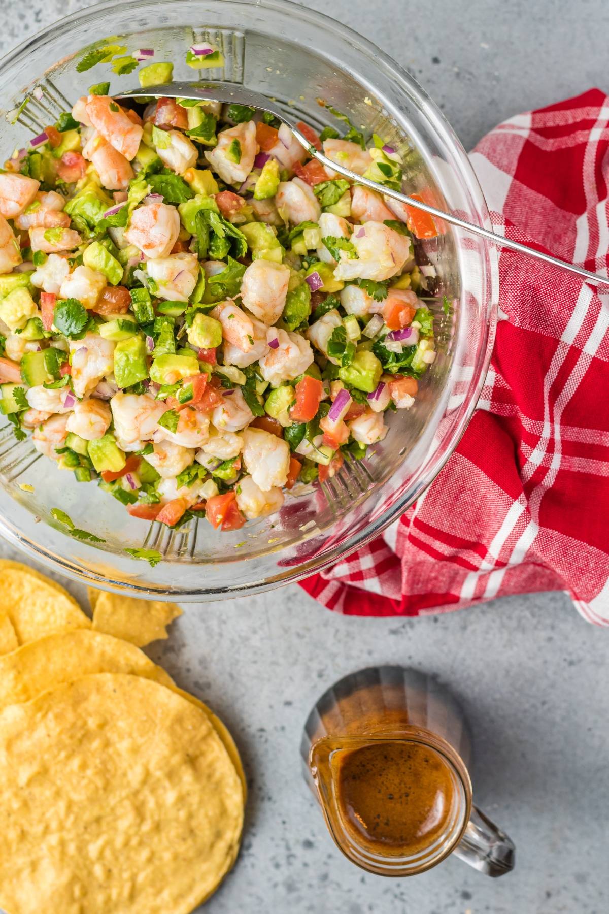 A glass bowl of shrimp ceviche, made with avocado, sits next to tostadas, a sauce, and a red checkered towel—perfect for anyone craving the best ceviche recipe.