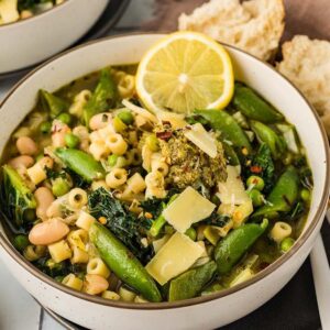 A bowl of minestrone verde, filled with pasta, beans, greens, snap peas, and a lemon slice, served alongside fresh bread.