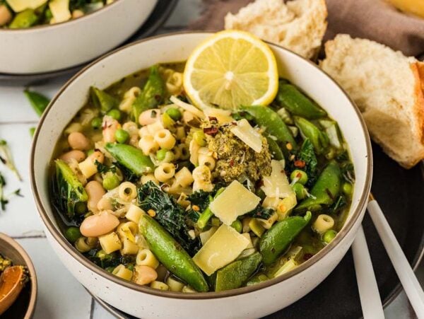 A bowl of minestrone verde, filled with pasta, beans, greens, snap peas, and a lemon slice, served alongside fresh bread.
